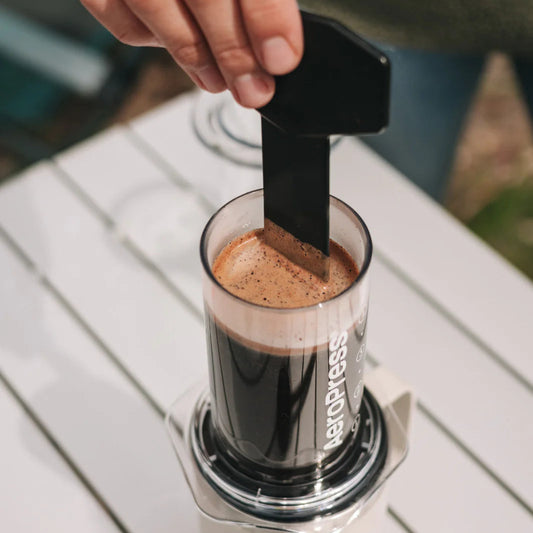 Person using a hand to press a black plunger into an AeroPress coffee maker on a white surface.