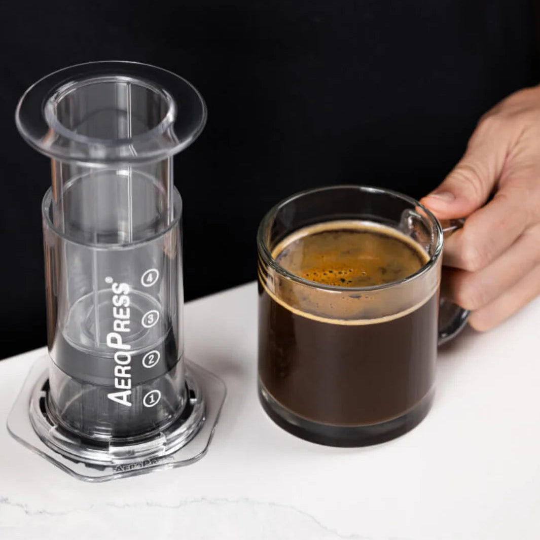 AeroPress coffee maker next to a glass of coffee on a white surface with a black background